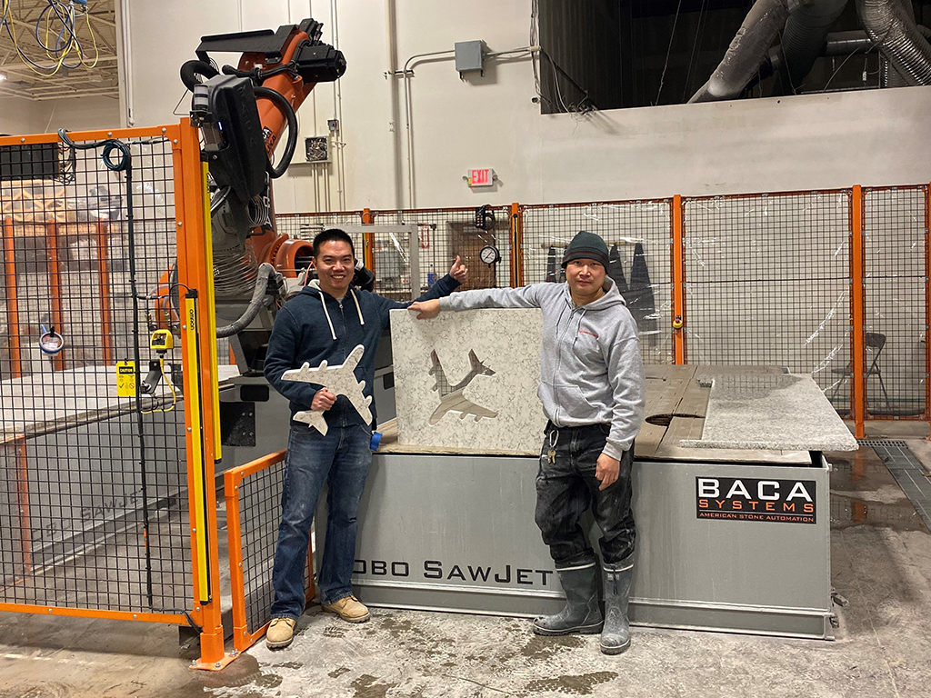 Two BACA employees standing in front of the Robo Sawjet. One employee has his arm on a stone slab that has an airplane engraved into it.